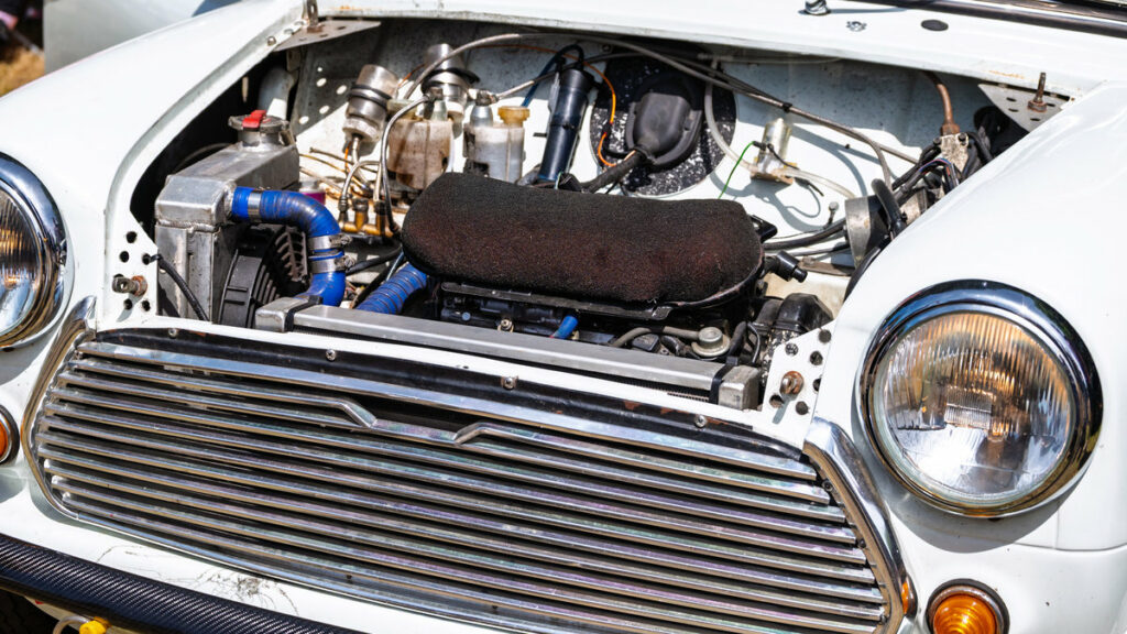 White car with open hood showing engine bay. Visible are a radiator, blue hoses, black air filter, front grille, headlights, and a carbon fiber bumper.
