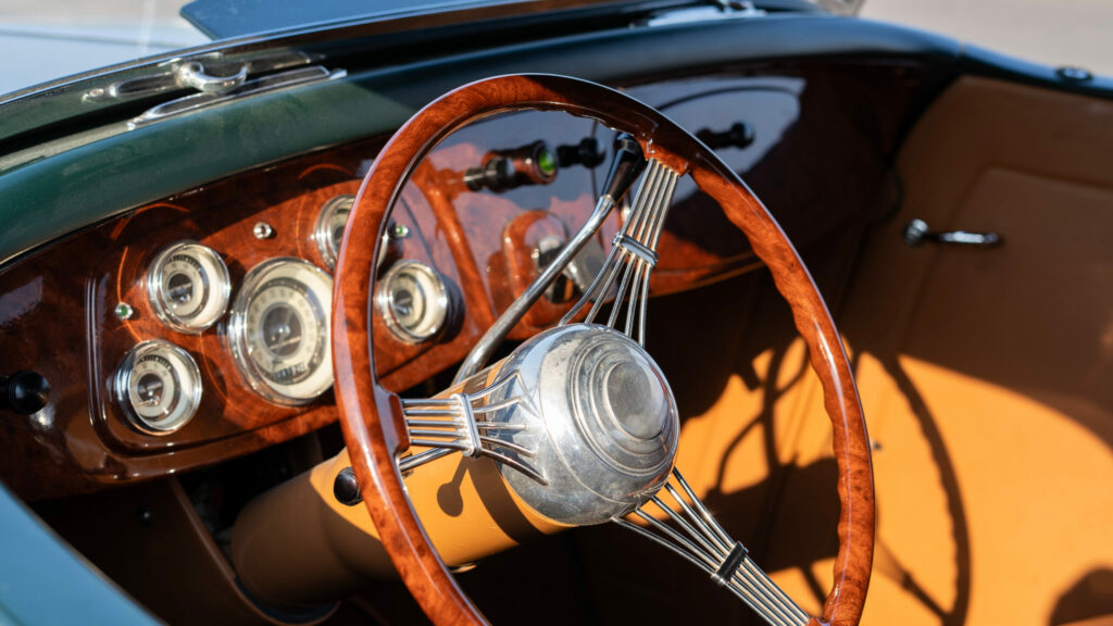 Close up of a classic vintage car interior featuring a polished wooden steering wheel, chrome details, analog gauges and elegant leather upholstery.